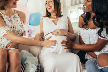 Gruppe von lächelnden jungen Freundinnen, die den Bauch einer schwangeren Frau berühren. Multirassische Freundinnen haben Spaß bei einer Babyparty. - JLPSF25344