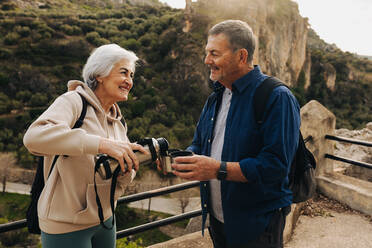 Cheerful senior couple taking a coffee break while backpacking outdoors. Happy elderly couple enjoying a leisurely hike in the hills. Adventurous couple spending quality time together after retirement. - JLPSF25349