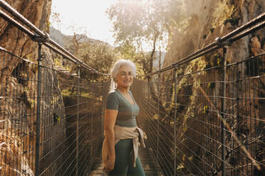 Glückliche ältere Frau, die in die Kamera lächelt, während sie auf einer Brücke im Freien steht. Fröhliche ältere Frau, die beim Wandern Musik hört. Reife Frau, die nach dem Ruhestand Freizeitaktivitäten genießt. - JLPSF25357