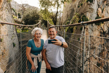 Lächelndes älteres Paar, das ein Selfie auf einer Brücke macht. Fröhliches älteres Paar, das eine gemütliche Wanderung im Freien genießt. Älteres Paar, das nach dem Eintritt in den Ruhestand gemeinsam glückliche Erinnerungen schafft. - JLPSF25359