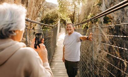 Ältere Frau, die ihren Mann auf einer Brücke fotografiert. Fröhliches älteres Paar, das eine gemütliche Wanderung im Freien genießt. Älteres Paar, das nach der Pensionierung gemeinsam glückliche Erinnerungen schafft. - JLPSF25360