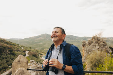 Happy senior man looking at the view while standing on a hilltop with binoculars. Cheerful elderly man enjoying a leisurely hike outdoors. Mature man enjoying recreational activities after retirement. - JLPSF25366