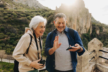 Happy elderly couple having a video call while standing on a hilltop. Cheerful senior couple enjoying a recreational hike outdoors. Adventurous couple having a good time together after retirement. - JLPSF25369