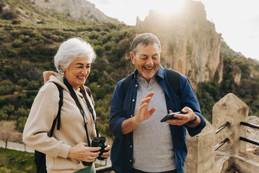 Mature couple having a video call while standing on a hilltop. Cheerful senior couple enjoying a recreational hike outdoors. Happy elderly couple having a good time together after retirement. - JLPSF25370