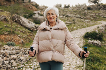 Glückliche ältere Frau, die in die Kamera lächelt, während sie auf einem Wanderweg mit Wanderstöcken steht. Abenteuerlustige ältere Frau, die eine gemütliche Wanderung genießt. Frau, die Freizeitaktivitäten nach der Pensionierung genießt. - JLPSF25371