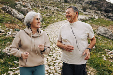 Happy senior couple smiling at each other while jogging down a hilly trail. Sporty senior couple listening to music while working out. Elderly couple maintaining a healthy lifestyle after retirement. - JLPSF25372