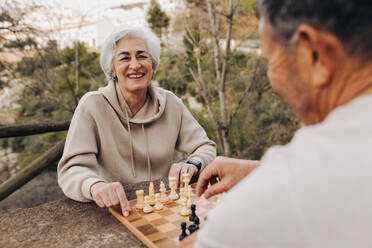 Mature couple playing a game of chess in a park. Cheerful senior couple spending some quality time together after retirement. Happy elderly couple having a good time outdoors. - JLPSF25374