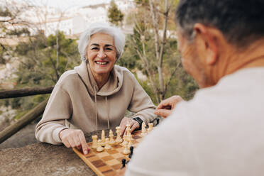 Fröhliches älteres Paar, das in einem Park Schach spielt. Fröhliches älteres Paar, das nach der Pensionierung viel Zeit miteinander verbringt. Reifes Paar, das sich im Freien vergnügt. - JLPSF25375