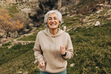 Happy senior woman smiling at the camera while jogging outdoors. Cheerful elderly woman listening to music while working out. Mature woman maintaining a healthy lifestyle after retirement. - JLPSF25377