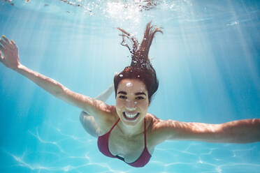 Unterwasseraufnahme einer Frau im Schwimmbad. Lächelnde Frau schwimmt unter Wasser im Pool eines Ferienorts. - JLPSF25381