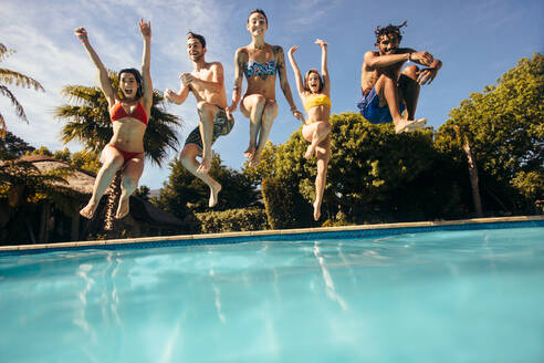 Fröhliche junge Freunde, die in einen Swimmingpool im Freien springen und Spaß haben. Gruppe von Männern und Frauen, die in den Pool eines Urlaubsortes springen. - JLPSF25384