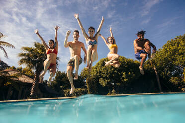 Group of friends jumping in swimming pool with their hands raised. Men and women jumping in pool and having fun. - JLPSF25387