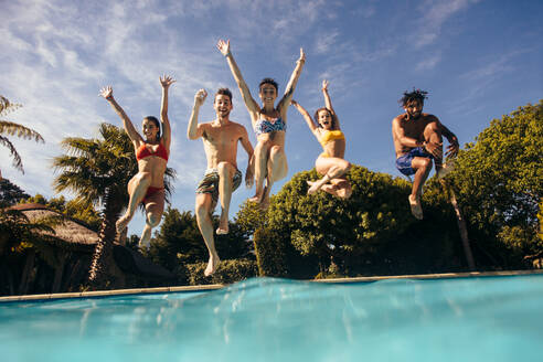 Eine Gruppe von Freunden springt mit erhobenen Händen in ein Schwimmbecken. Männer und Frauen springen in ein Schwimmbecken und haben Spaß. - JLPSF25387