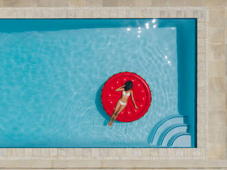 Draufsicht auf eine Frau, die sich auf einer aufblasbaren Matratze im Schwimmbad entspannt und ihr Gesicht mit einem Hut bedeckt. Frau im Bikini sonnt sich auf aufblasbarer Matratze im Schwimmbad. - JLPSF25400