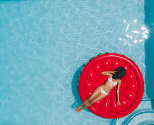 Draufsicht auf eine Frau im Bikini, die sich auf einer schwimmenden Matratze im Poolwasser an einem heißen sonnigen Tag entspannt. - JLPSF25401