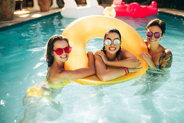 Porträt von fröhlichen Frauen im Pool mit Sonnenbrille und aufblasbarem Ring. Gruppe von glücklichen Freundinnen, die den Sommer im Pool genießen. - JLPSF25416