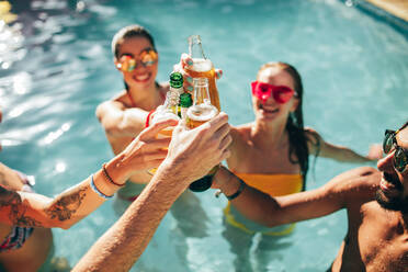 Fröhliche Gruppe junger Leute im Schwimmbad, die ein Bier trinken. Gemischtrassige Freunde, die bei einer Poolparty Getränke genießen und darauf anstoßen. - JLPSF25431