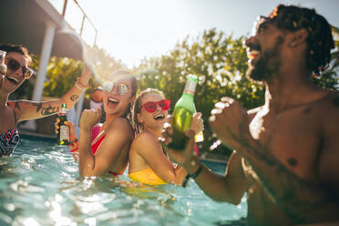 Group of happy friends at pool party with beers. Men and women people laughing and having fun inside swimming pool. - JLPSF25434