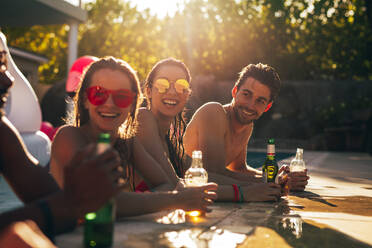 Eine Gruppe junger Leute steht am Rande des Swimmingpools mit Bier auf einer Sommerparty. Männer und Frauen im Swimmingpool lachen und reden auf einer Poolparty. - JLPSF25435