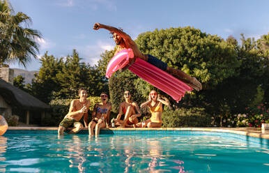 Man jumping in the swimming pool with inflatable mattress and friends sitting on the edge of the pool. Friends enjoying a summer day at pool side. - JLPSF25438