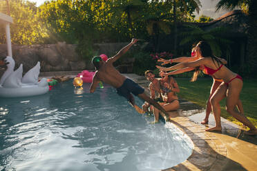 Group of friends enjoy pool party together in summertime. Women pushing male friends in the swimming pool. - JLPSF25440