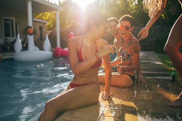 Frauen, die am Rande des Pools sitzen, sich unterhalten und lächeln. Freunde feiern eine Poolparty an einem Sommertag. - JLPSF25442