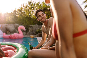 Handsome young man sitting on the edge of swimming pool with female friends. Young people having pool party during summer holiday. - JLPSF25445