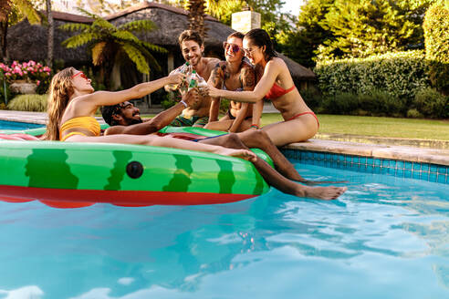 Eine gemischtrassige Gruppe von Freunden feiert in einer privaten Villa am Pool. Männer und Frauen stoßen auf einer Poolparty mit Bier an. - JLPSF25447
