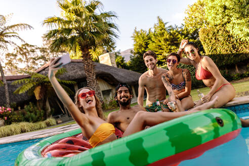 Verrückte Menschen, die den Sommer am Pool genießen und ein Selfie machen. Frau sitzt auf einer aufblasbaren Matratze und macht ein Selfie mit Freunden bei einer Poolparty. - JLPSF25452