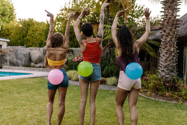 Rear view of three women standing outdoors with balloons tied on their back. Friends playing balloon bursting game at party. - JLPSF25456