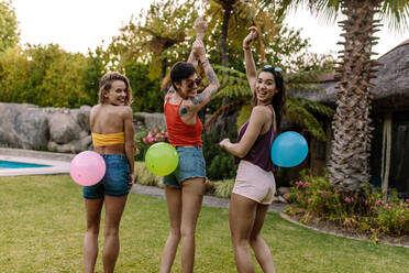 Lächelnde Frauen stehen im Freien mit Luftballons auf dem Rücken gebunden. Gruppe von Freunden genießen Ballon Pop Spiel auf der Party. - JLPSF25457