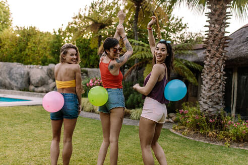 Lächelnde Frauen stehen im Freien mit Luftballons auf dem Rücken gebunden. Gruppe von Freunden genießen Ballon Pop Spiel auf der Party. - JLPSF25457