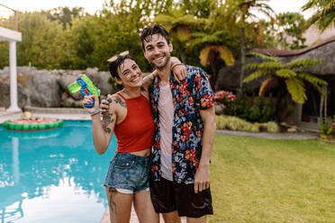 Cheerful young couple standing by the pool with water gun. Woman holding water gun standing with her boyfriend. Both fully wet with water. - JLPSF25462