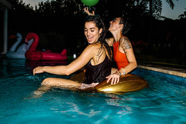 Glückliche Frauen im Pool mit aufblasbarem Ring. Weibliche Freunde haben Spaß während einer abendlichen Poolparty. - JLPSF25463