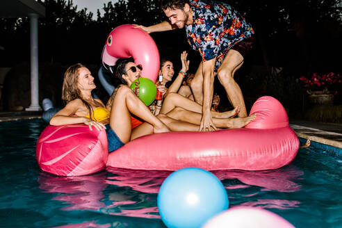 Fröhliche Männer und Frauen im Schwimmbad auf einem aufblasbaren Schwimmer. Gruppe von glücklichen Freunden, die abends in einem Schwimmbad feiern. - JLPSF25470