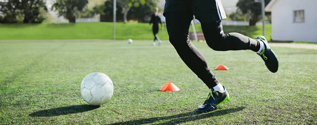 Niedriger Blickwinkel eines jungen Fußballspielers, der auf einem Fußballfeld rennt und den Ball zwischen einer Reihe von Kegeln hindurchführt, während des Trainings. - JLPSF25498