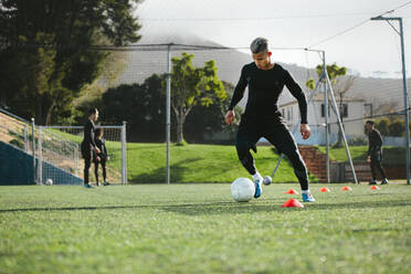 Junger Fußballspieler, der auf einem Fußballplatz trainiert, mit einer Mannschaft im Hintergrund. Fünf Fußballspieler trainieren auf einem Feld im Freien. - JLPSF25506