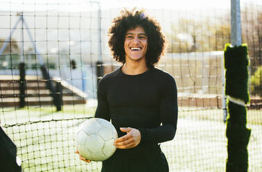 Portrait of teenage football player holding a ball in his hand and laughing. Young man training on soccer field. - JLPSF25515