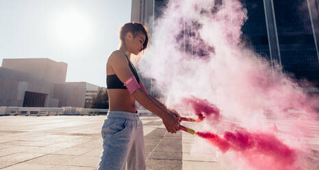 Sporty young woman holding smoke sticks outdoors in city. Sportswoman holding a smoke grenades over urban city background. - JLPSF25535