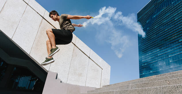 Free runner jumping over an obstacle with smoke grenades. Young sportsman doing parkour stunts outdoors in the city. - JLPSF25541