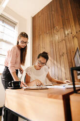 Vertikale Aufnahme von zwei Geschäftsfrauen, die gemeinsam an einem neuen Projekt arbeiten. Eine Mitarbeiterin geht an ihrem Schreibtisch Papierkram durch. - JLPSF25560