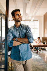 Porträt eines jungen Geschäftsmannes, der an der Tür steht, während im Hintergrund Kollegen arbeiten. Junger Mann in einem Startup-Büro. - JLPSF25570