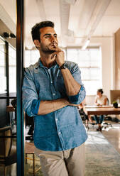 Portrait of young man standing at door looking away and thinking. Thoughtful young male at startup office with colleagues working in background. - JLPSF25571