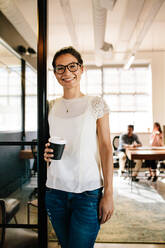 Portrait of smiling young woman standing in office doorway with coffee. Relaxed female executive having coffee break with colleagues working in background. - JLPSF25574