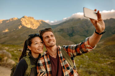Junges Paar beim Wandern nimmt Selfie mit Smartphone. Glücklicher junger Mann und Frau nehmen Selbstporträt mit Berglandschaft im Hintergrund. - JLPSF25591