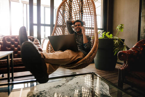 Glückliche junge Geschäftsmann sitzt auf einem Korb Hängesessel im Büro Lounge mit Laptop und sprechen auf dem Handy. Männliche Führungskraft entspannt im Büro Lounge während der Pause. - JLPSF25602