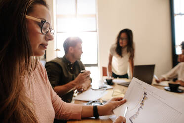 Close up of young woman looking at document during meeting in conference room. Businesswoman reading business chart with people in background. - JLPSF25609