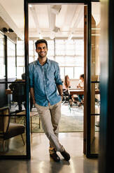 Full length portrait of handsome young man standing in doorway of office with his hands in pockets. Creative male executive at startup with colleagues in background. - JLPSF25637