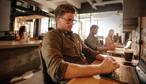 Young man sitting at cafe and using laptop computer, with people in background. - JLPSF25651