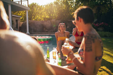 Außenaufnahme einer Gruppe junger Leute, die am Rande des Pools sitzen und Bier trinken. Multirassische Freunde genießen und trinken während einer Poolparty. - JLPSF25658